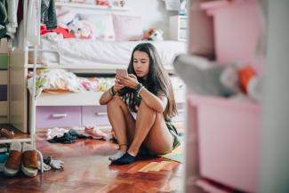 A teenage girl sits on her bedroom floor, texting with her mobile phone